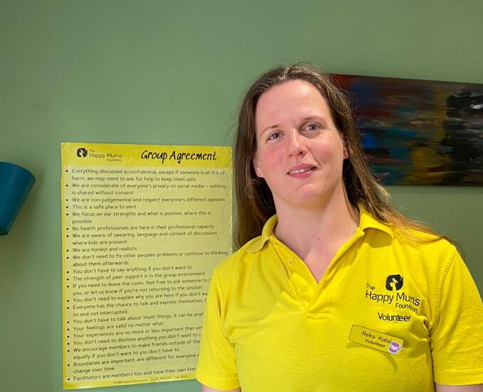 A Happy mums volunteer in a statement yellow polo shirt standing in front of a green wall with a yellow Group Agreement Poster.